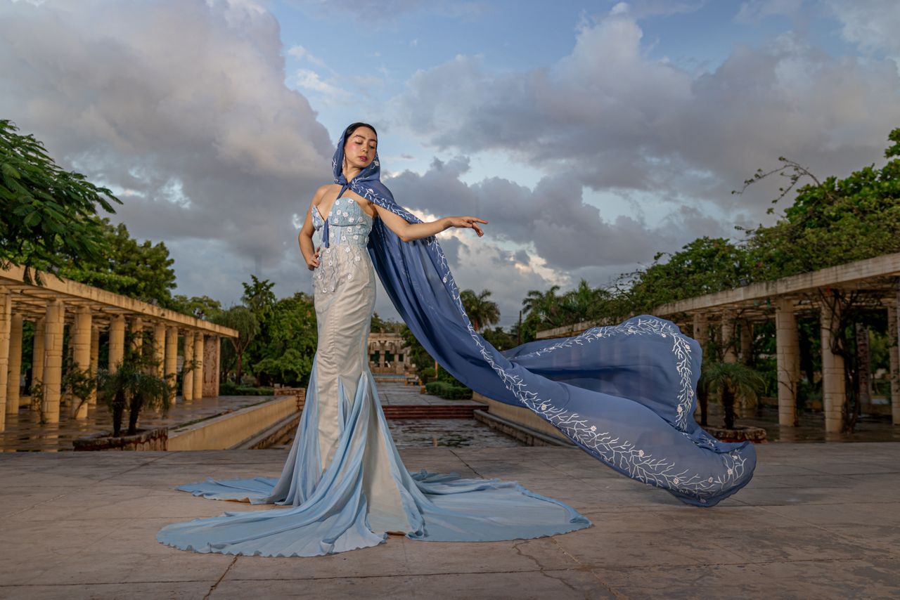 about-06 Elegant woman in a detailed blue dress poses gracefully outdoors with dramatic sky.